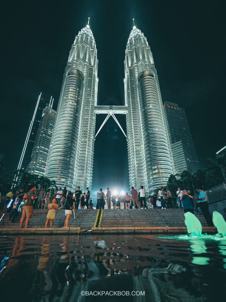 Tourists are visiting the Petronas Towers in Kuala Lumpur for free. The photo shows both towers and the water feature at the bottom the is a crowd of tourists taking free photos