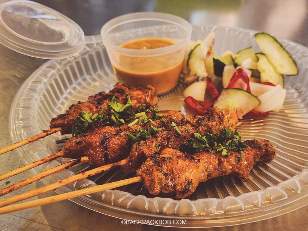 A plate of chicken satay skewers at the free to visit Kuala Lumpur Jalan Alor Market. There is a clear plastic plate with 5 meat skewers and a vegetable garnish, there is a tub or peanut sauce
