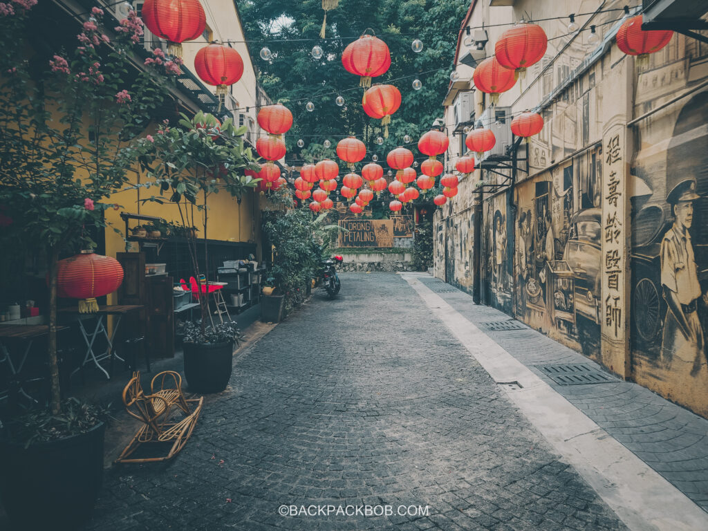 A street in Kuala Lumpur which is free to walk down. There are many chinese laterns hung between two buildings and the building have yellow walls painted with street art