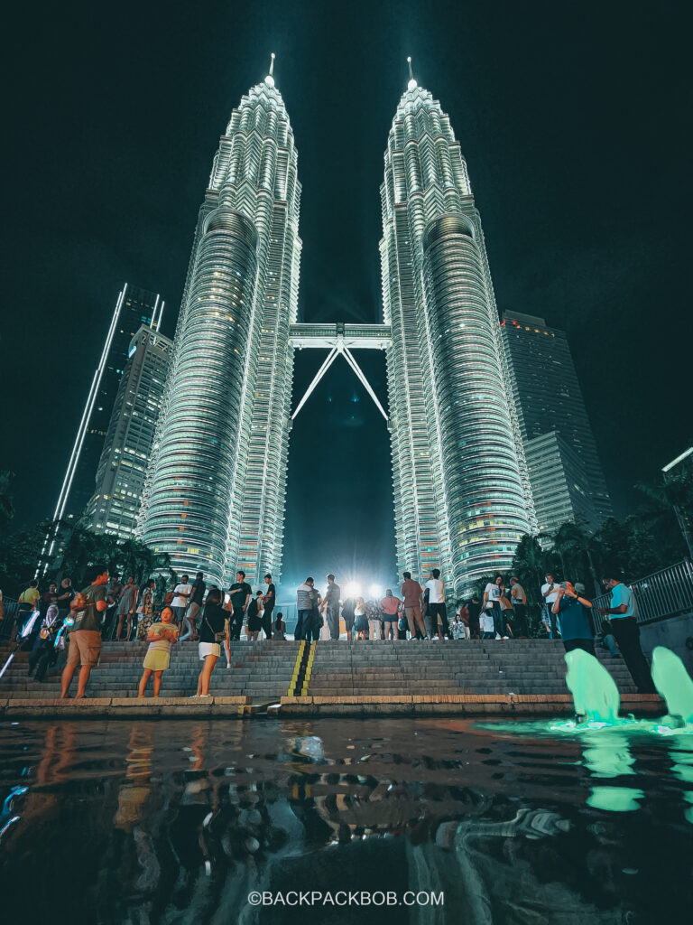 Tourists are visiting the Petronas Towers in Kuala Lumpur for free. The photo shows both towers and the water feature at the bottom the is a crowd of tourists taking free photos 