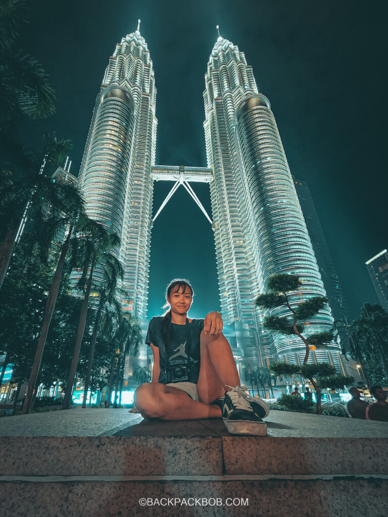 The Kuala Lumpur Petronas Towers are iluminated in the evening on Malaysia itinerary Kuala Lumpur backpack bob is sat in front of the towers which are blue