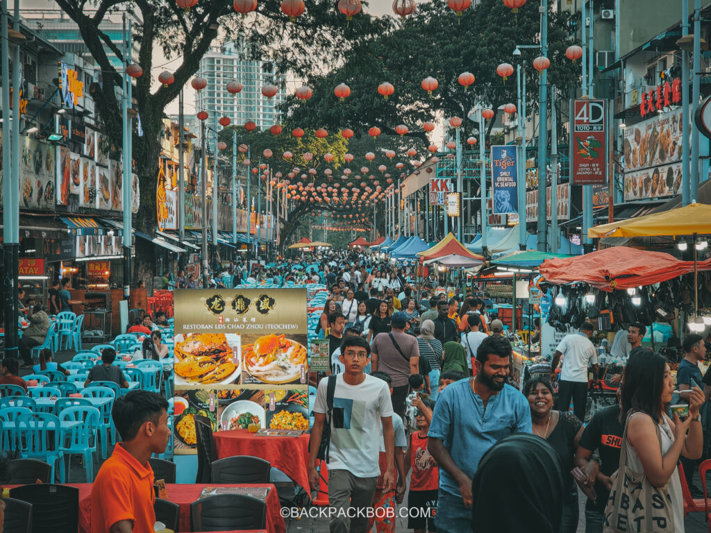 Jalan Alor Street Market is free to visit in Kuala Lumpur there are crowds of people in the photo walking in the street, on either side of the street there are plastic tables and chairs to sit and eat