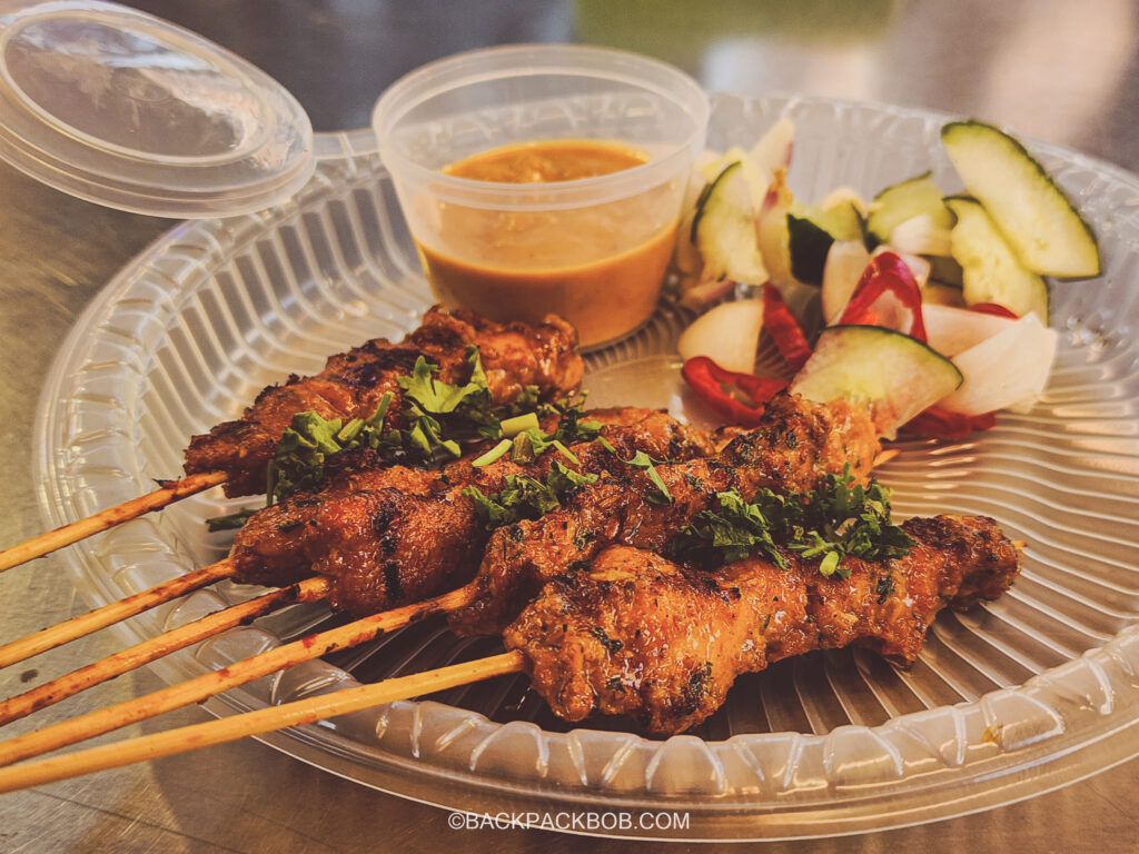 A plate of chicken satay skewers at the free to visit Kuala Lumpur Jalan Alor Market. There is a clear plastic plate with 5 meat skewers and a vegetable garnish, there is a tub or peanut sauce